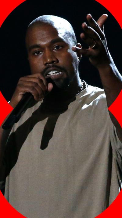 Kanye West on stage holding a microphone with his right hand whilst his left hand is held up. He looks past the camera and wears a green t-shirt.