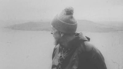 Man wearing rain gear and bobble hat looks out at an island in the distance