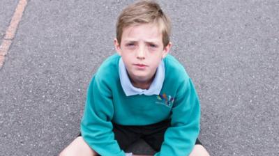 Young schoolboy sits on playground tarmac looking at the camera