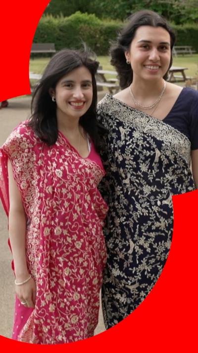 Two young women, both with long dark hair, stand side by side smiling. They are both wearing intricately-patterned saris.