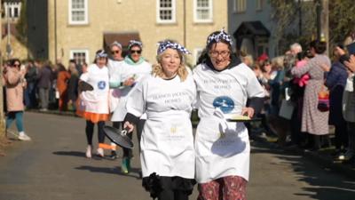 Olney Pancake Race - two women running the three-legged race wearing white aprons, chequered headscarves and carrying frying pans in a crowd-lined street