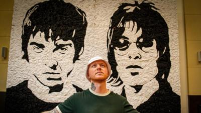 The artist standing with his arms outstretched at though on the cross with the full mural in the background. He is dressed the same, but has on a white bucket hat.