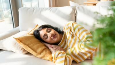 A woman naps. She has black hair and a yellow and white striped top. Sun streams in the window.
