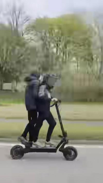 Two youths on an e-scooter on a public road with trees behind.
