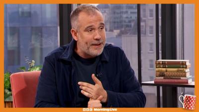 Dr Xand in the Morning Live studio sat down on an orange chair, wearing a navy blue shirt.