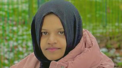 A woman in her 20s wearing a black hijab and pink puffy coat. There is grass behind her although the background is blurred. She has a slightly smiling but fairly neutral expression as she looks at the camera.
