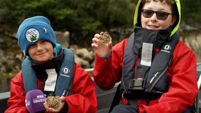 Two boys wearing life jackets and waterproofs sit on a boat, smiling, holding up oysters to the camera