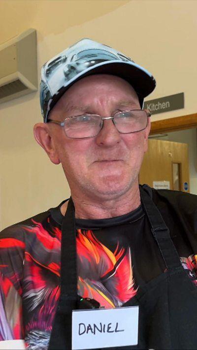 A man wearing an apron and a baseball cap stands in a community kitchen