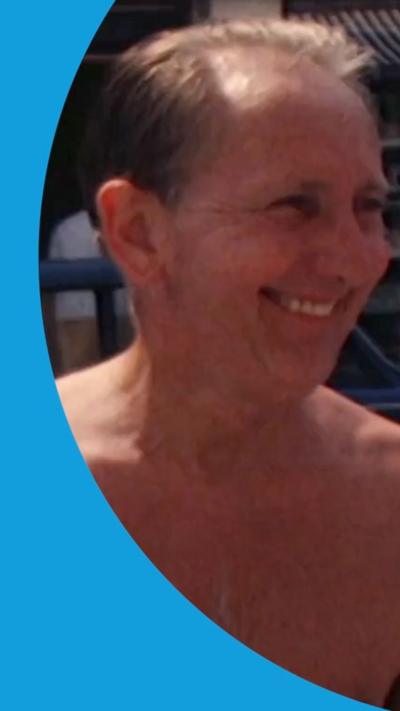A man that has his top off in the sun. he is smiling at someone off camera. He has short dark hair.