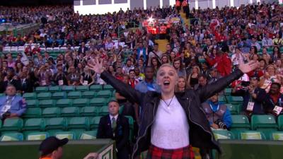 Comedian Karen Dunbar stands in front of a crowd of spectators wearing a red kilt and black leather jacket. 