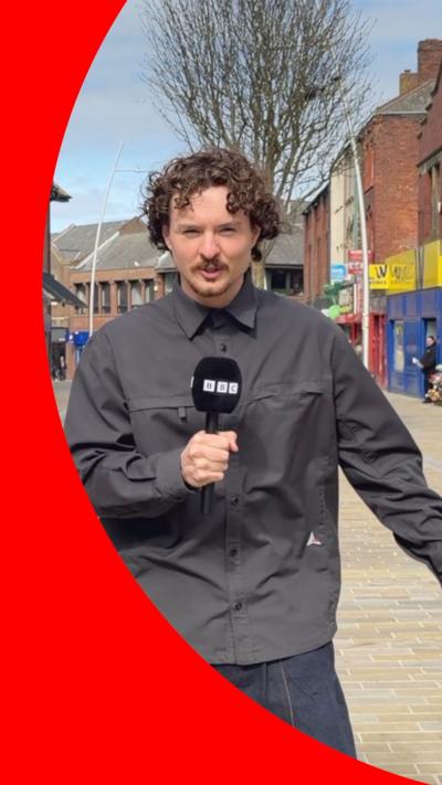 BBC reporter Toby Bewley stands holding a microphone in Barrow Town Centre.