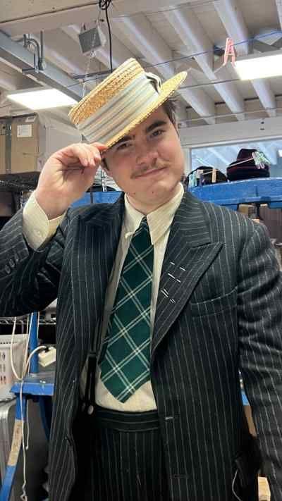 A young man wearing a pinstripe suit and a checked green and blue tie. He wears a straw hat, which with his right hand, he is doffing towards the camera. He stands inside.