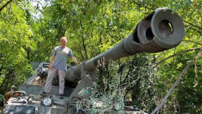 Jack Lopresti dressed in grey T-shirt and trousers stood on huge tank surrounded with trees when he previously served in the UK Army Reserve as a corporal