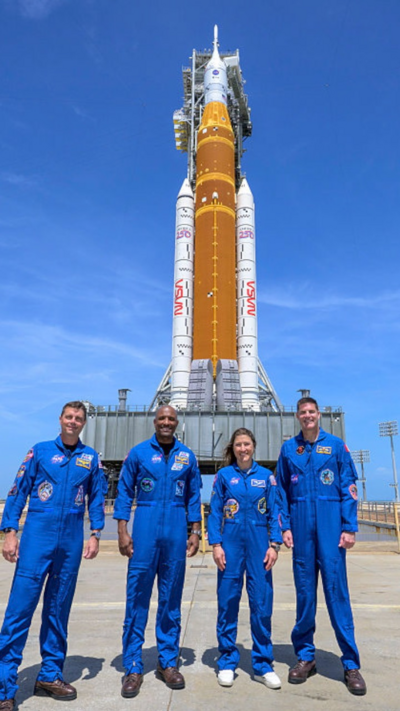 Four astronauts in blue overalls stand on the tarmac in front of the rocket carrying the Orion spacecraft