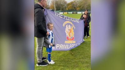 Two adults and two children stand on a football field holding a flag for Wythenshawe AFC