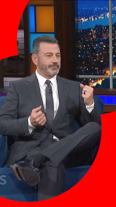 Jimmy Kimmel sits on the Stephen Colbert set in a black suit with a black tie