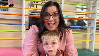 A woman wearing pink with brown hair and glasses holds a little blonde haired boy with pink glasses in her arms 
