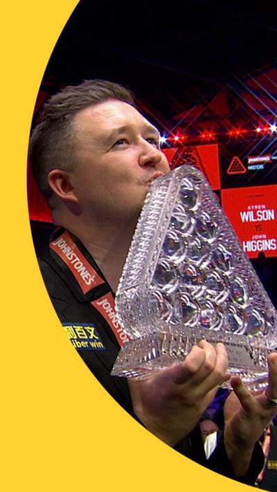 A man in a black shirt kisses a large glass trophy