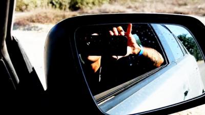 A man holds his phone up to car mirror