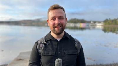 Jamie McDonald holds a microphone, behind him is a loch on the Isle of Skye 