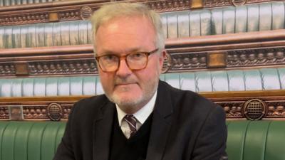 A white man with white hair in a suit looking at the camera outside of the House of Parliament in London