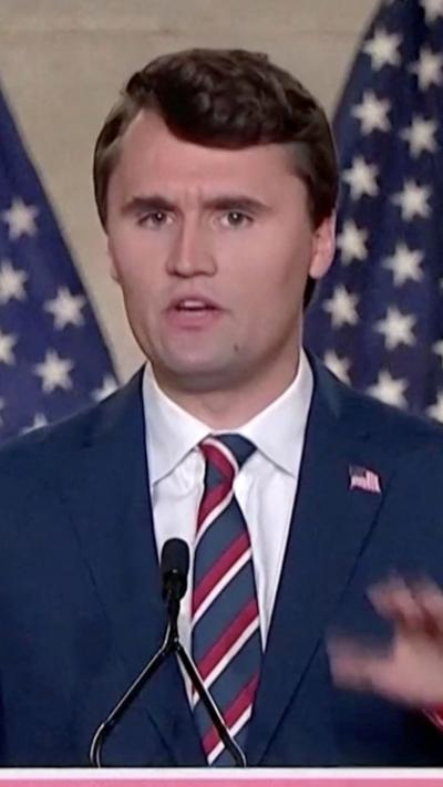 Charlie Kirk standing at podium with two USA flags waving behind him