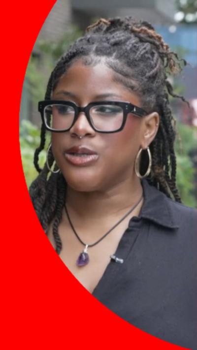 Woman with dreadlocks, black shirt, and purple necklace standing on a sidewalk near a brick building and greenery.