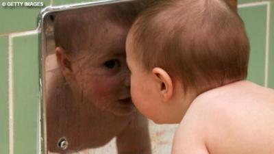Image of baby leaning its head against a mirror. The baby is looking away from the camera but the reflection image is looking back towards the camera. There are green tiles around the mirror.