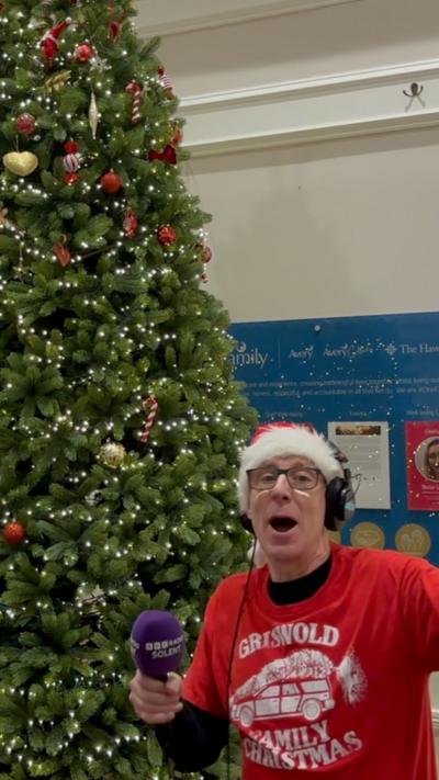 A man in a Santa hat and a red t-shirt standing next to a Christmas tree