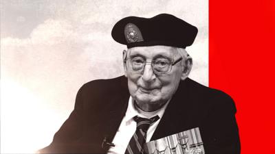 An older man looking directly into the camera. He is wearing a shirt, tie and a jacket which has medals on the front. He is also wearing a beret and glasses.  