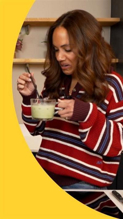 Alex Scott standing in a kitchen holding glass mug of matcha. Alex grimaces.