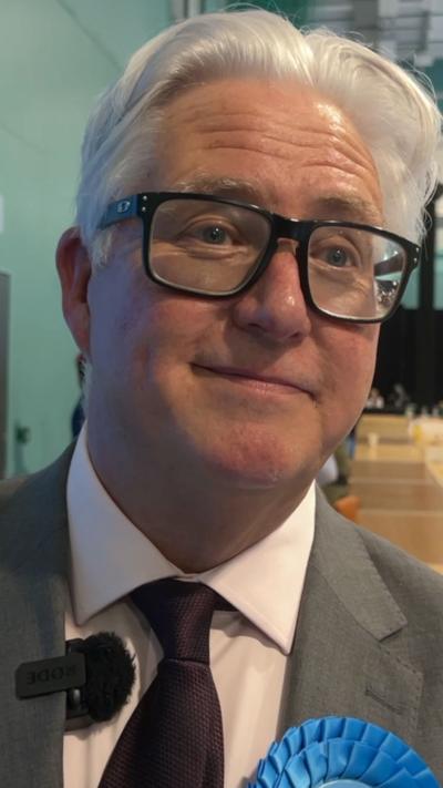 Kevin Bentley, wearing a grey suit and big square frame black glasses.