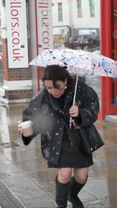 Two women stand on a rainy sidewalk, one holding a patterned umbrella and coffee.