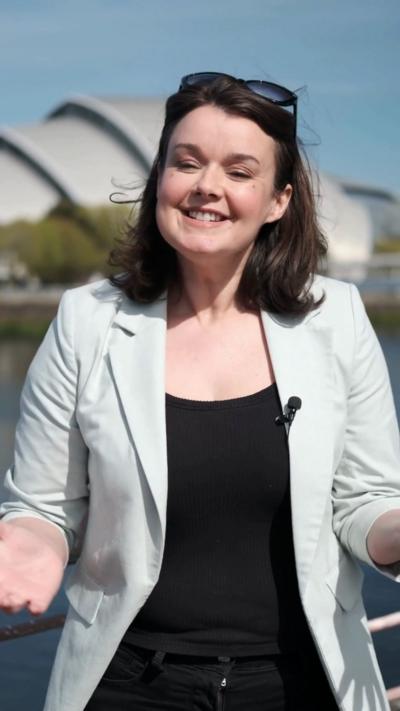 A woman with a brown bob, light blue blazer and sunglasses smiles at the camera. The SEC Armadillo is in the background.