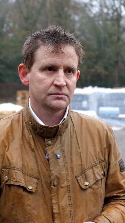A man in a brown coat. There are crates of bottled water behind him.