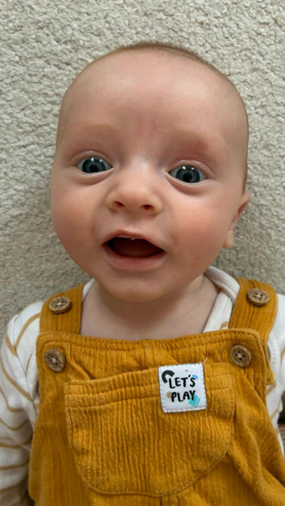 Baby Macsen lying on a pale carpet with yellow dungarees on that say let's play