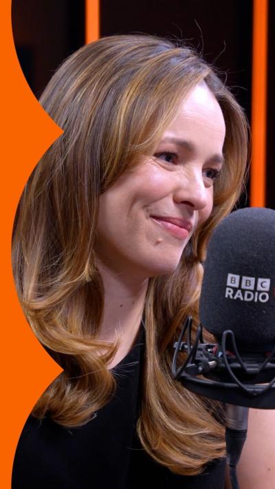 Rachel McAdams sits behind a BBC microphone, she has shoulder-length dark blonde hair and wears a black T-shirt