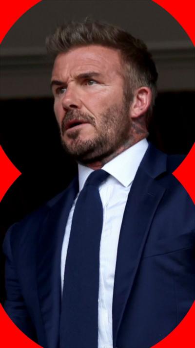 Sir David Beckham has short dark hair, a beard and a moustache. He is wearing a dark blue suit and tie, with a white shirt undernea