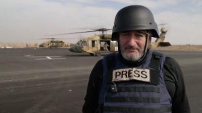 Fergal Keane wearing a helmet and a bullet proof vest that reads 'press', he is stood on an airfield in front of a helicopter