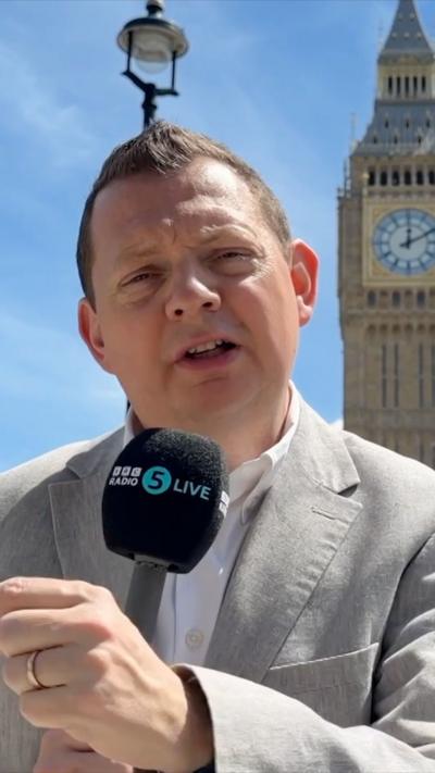 Matt Chorley is wearing a light grey suit with a white shirt. He is holding a microphone.