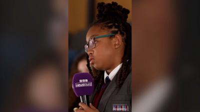 A girls with black braids and glasses holds a purple microphone