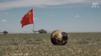 Lunar probe in a field, next to Chinese flag and with helicopter in bacground