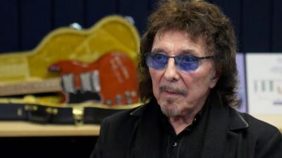 Man with blue-tinted glasses, beard and dark hair in a black jacket with black shirt. Behind him is a red guitar