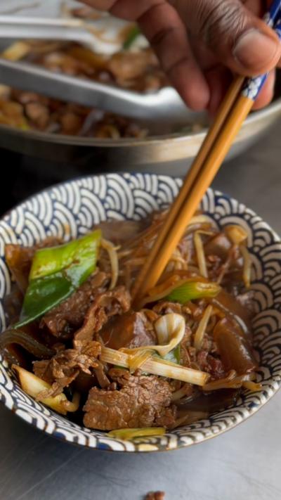 A hand holding chopsticks picks up noodles from a bowl of beef chow fun, on a table. A wok is in the background