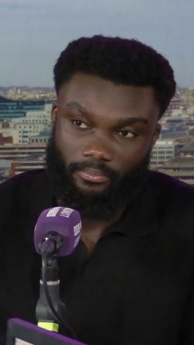 A person seated at a microphone, discussing against a backdrop of a city skyline.