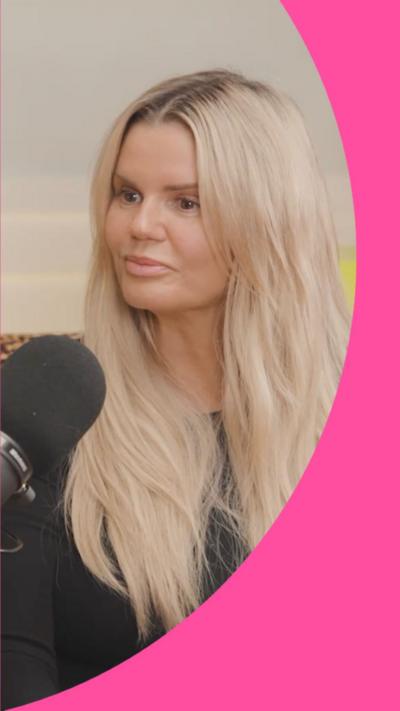 Kerry Katona has long blonde hair and is wearing a black top. She is sitting in front of a black microphone.