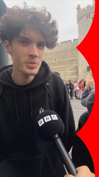 A young man in a black hoodie with wavy brown hair speaks into a BBC mic, outside Windsor Castle 