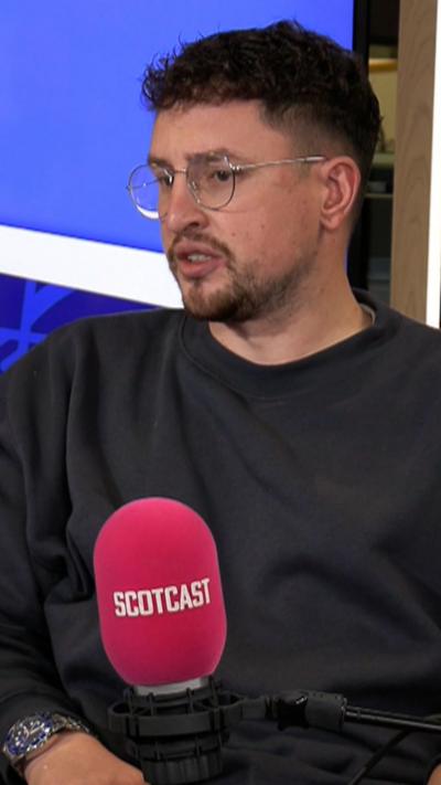 Jamie is wearing a dark jumper, silver round glasses and a silver watch. He is talking into a pink mic with the word 'Scotcast' on it.
