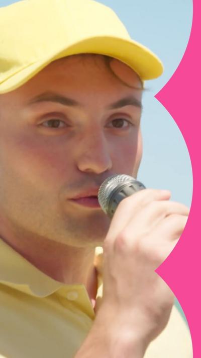 A man speaks into a mic. He wears a yellow baseball cap and T-shirt.