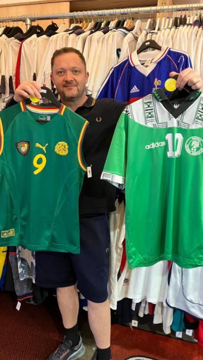 A man stands in front of a rack full of England football shirts holding up two football shirts. The shirt on the left is a 2002 Cameroon shirt which is sleevless. It is dark green with a yellow '9' on it. On the right is the 1994 World Cup shirt for Nigeria, it is mostly a light green with a white upper panel with some dark green patterns. 